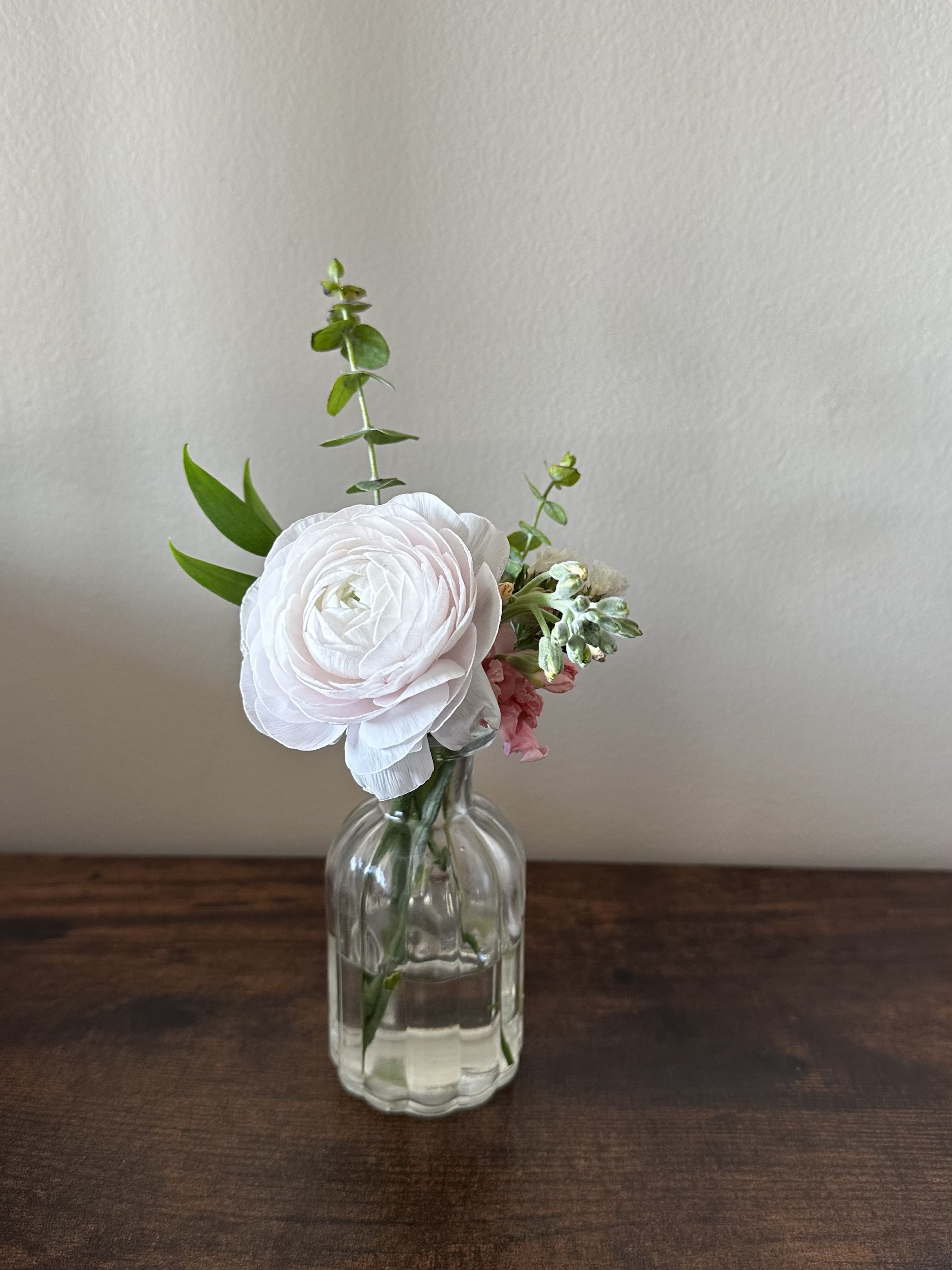 A small glass bud vase with pink and white flowers and green foliage on a wooden surface against a plain wall for wedding decor.