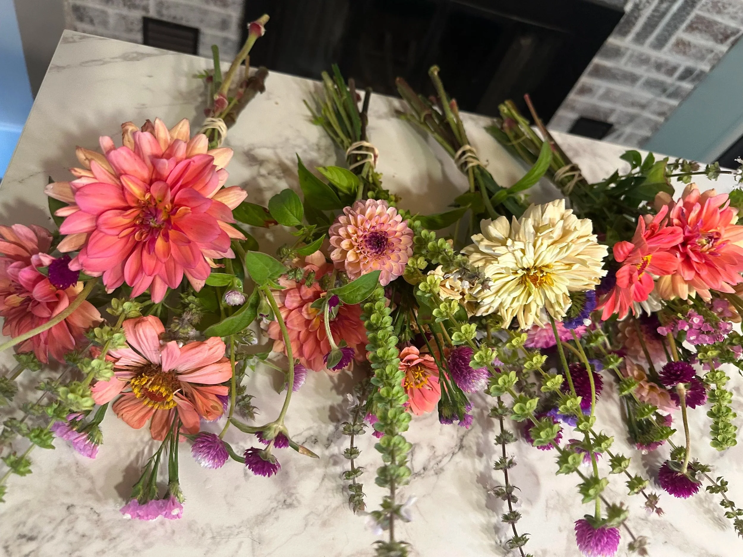 Colorful bouquet of various flowers including pink dahlias, cream-colored dahlia, and small purple and pink blossoms on a marble surface in front of a fireplace.