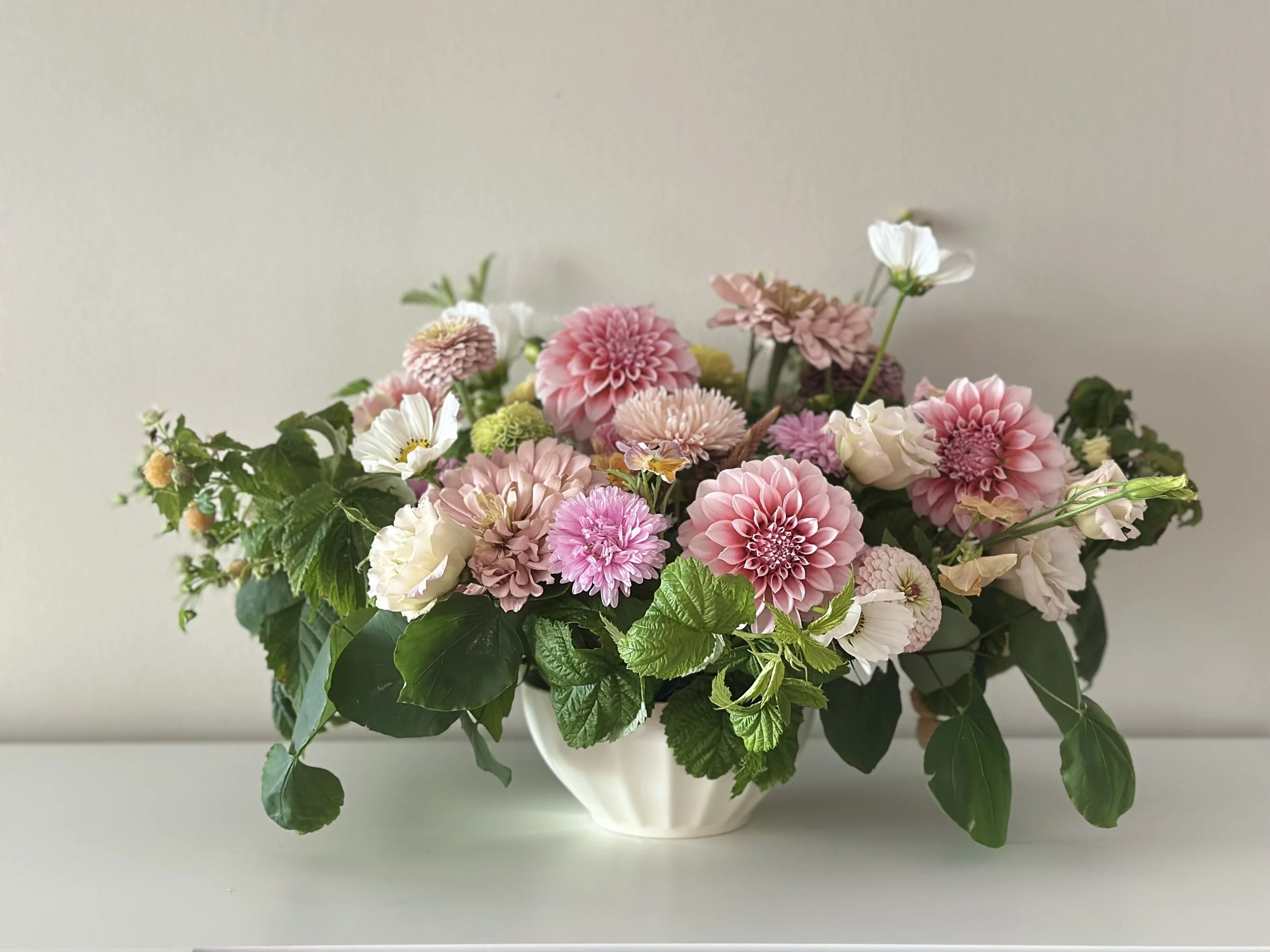 A white compote centerpiece filled with pink, white, and light purple dahlias and fresh local flowers and green leaves, placed on a white surface against a plain light-colored wall.