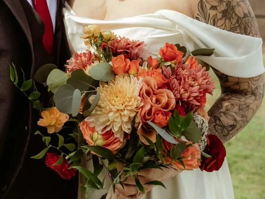 Person holding a bouquet of orange, peach, and white flowers with green leaves.