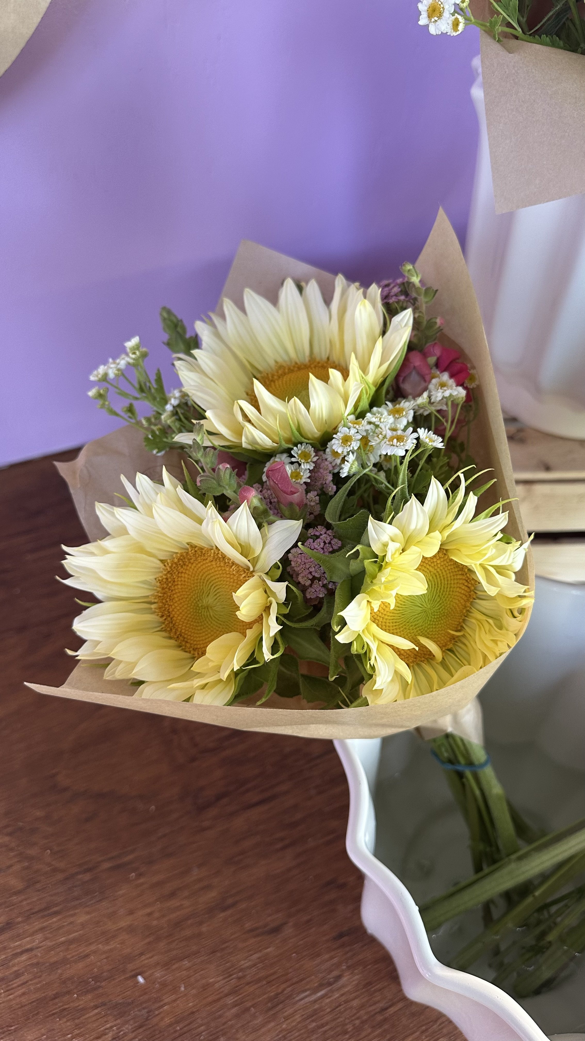 A bouquet of sunflowers, white daisies, pink flowers, and greenery wrapped in brown paper and set in a white vase.
