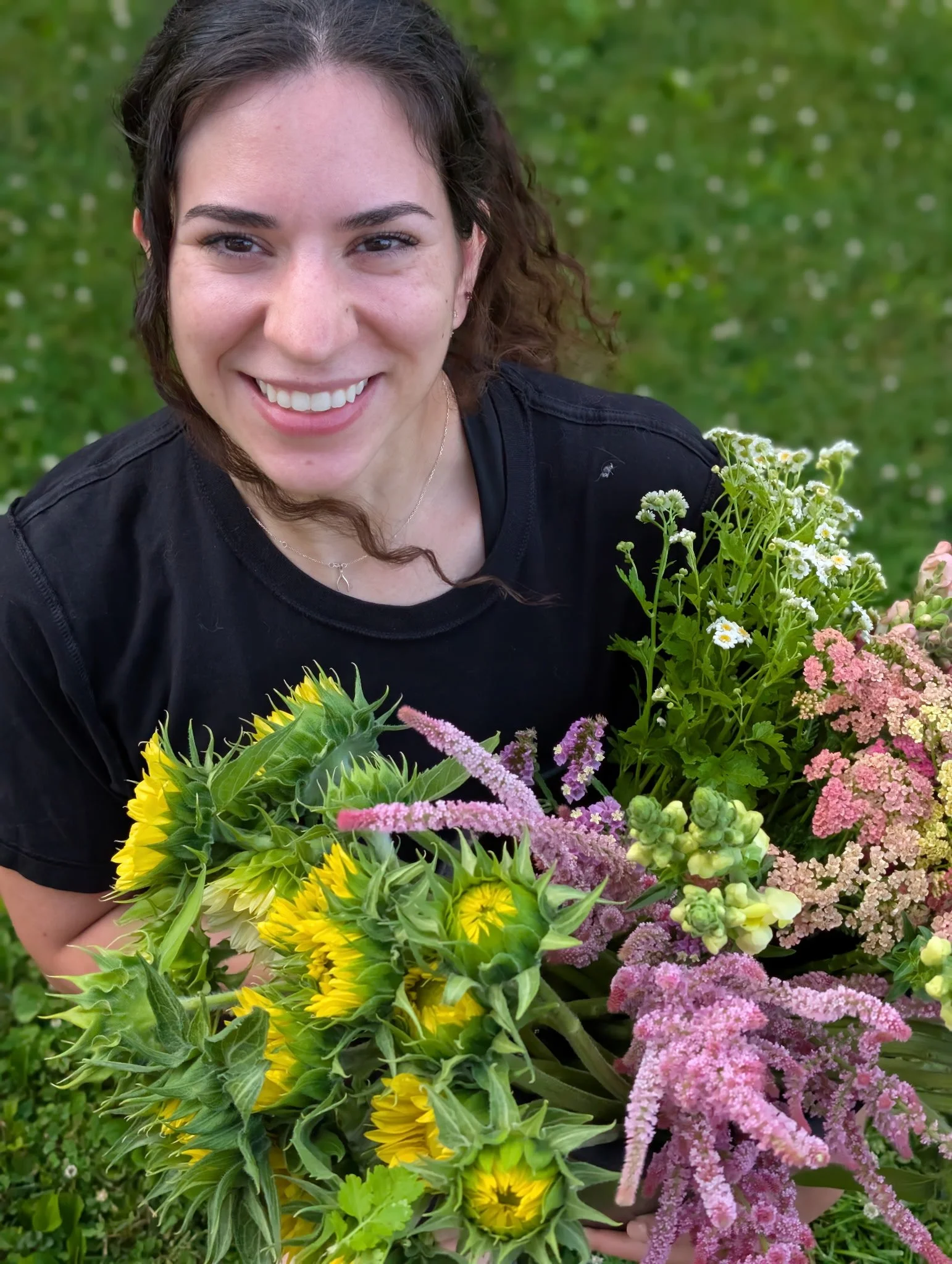 A woman smiling outdoors holding a large bouquet of colorful flowers including sunflowers, pink, white, and purple blooms, with a grassy background.