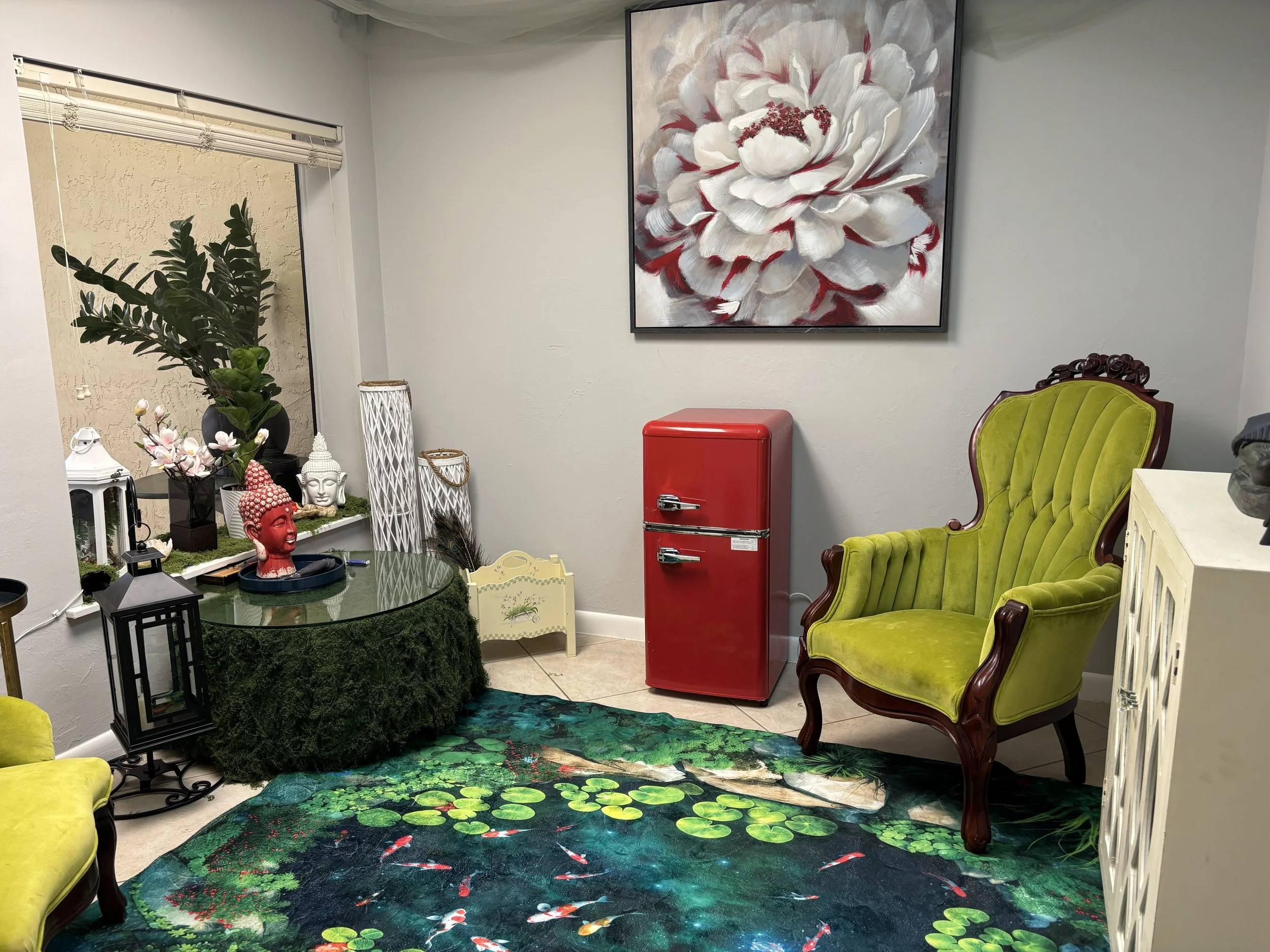 Decorative room with a large framed floral painting on the wall, a red vintage mini fridge, a lime green velvet armchair, various plants and statues, and a colorful lily pond rug on the floor.