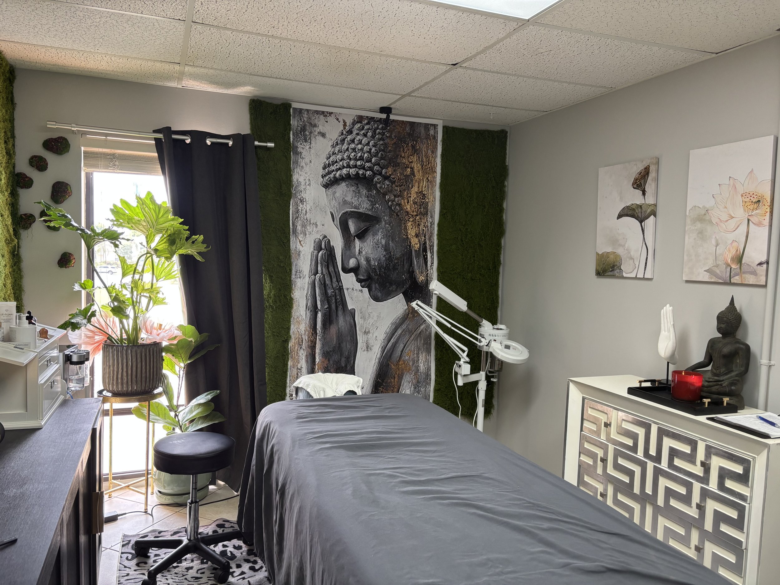A room with a massage or treatment bed covered with a gray sheet, a large Buddha painting on the wall, decorative moss and rocks on a shelf, a plant on a small table, artwork on the wall, and a window with black curtains.