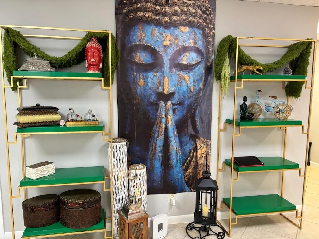Decorative shelves with green surfaces and gold frames displaying various objects, including a red Buddha head, woven baskets, a bicycle model, a small statue, and glass jars. A large portrait of a peaceful, clasped hands Buddha dominates the center,