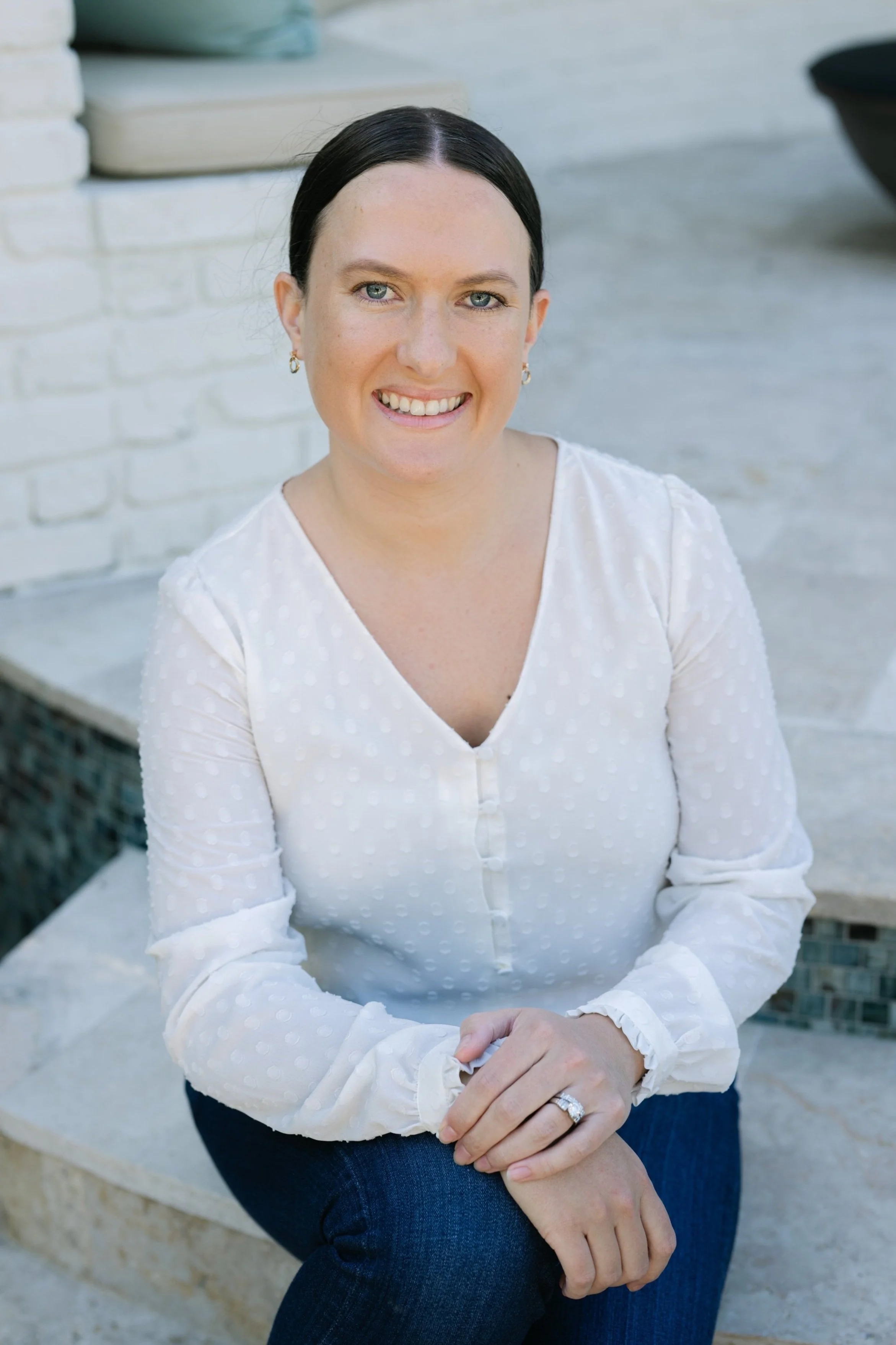 A woman with dark hair in a white blouse and dark pants sitting outdoors, smiling at the camera.