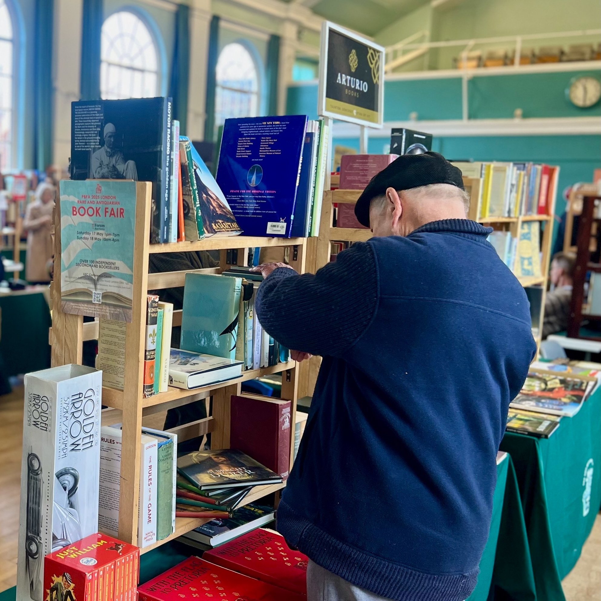Person browsing a bookshelf at a book fair, with various books and a sign for Arturo Books.