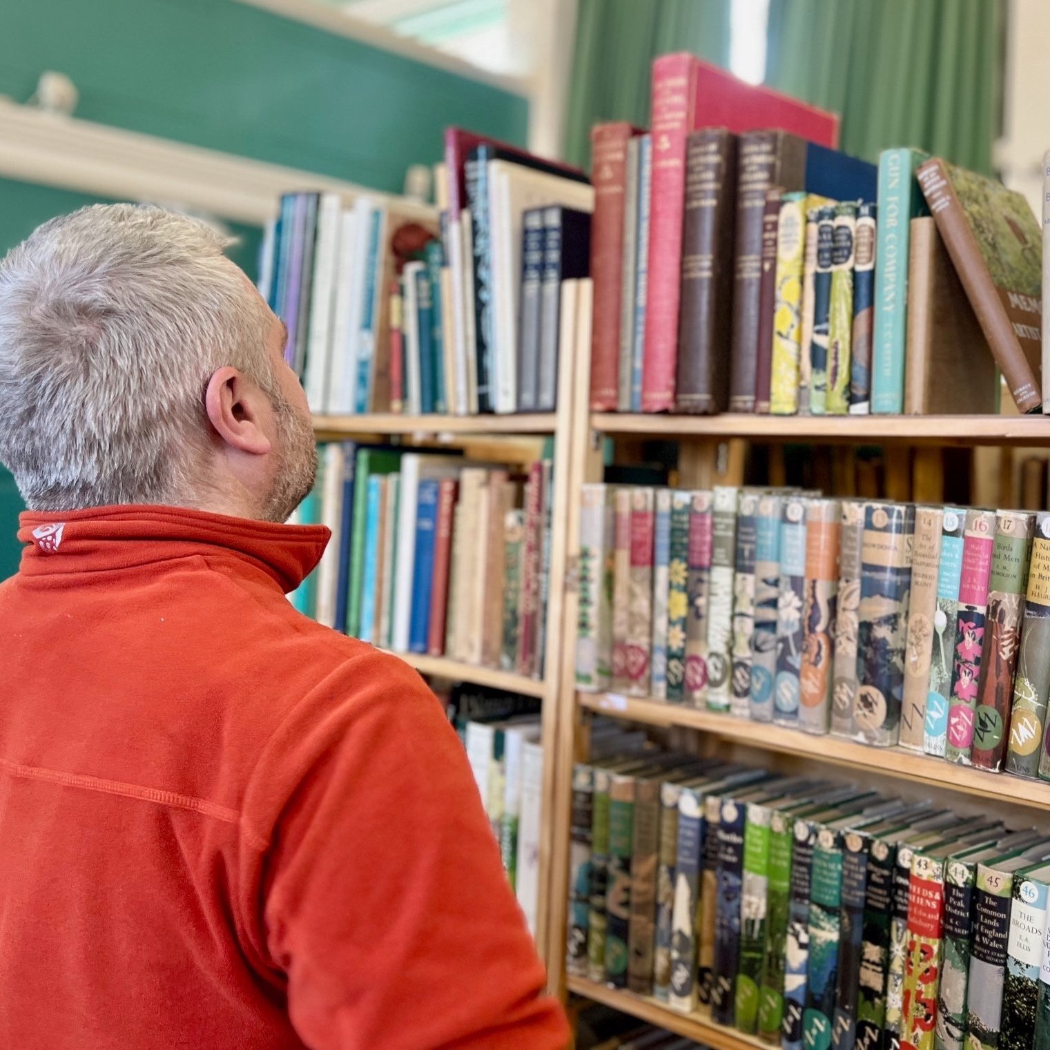 Person in orange jacket browsing books on a library shelf filled with various colorful hardcover books.
