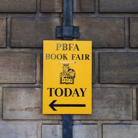 Yellow sign for PBFA Book Fair with arrow pointing left, mounted on brick wall.