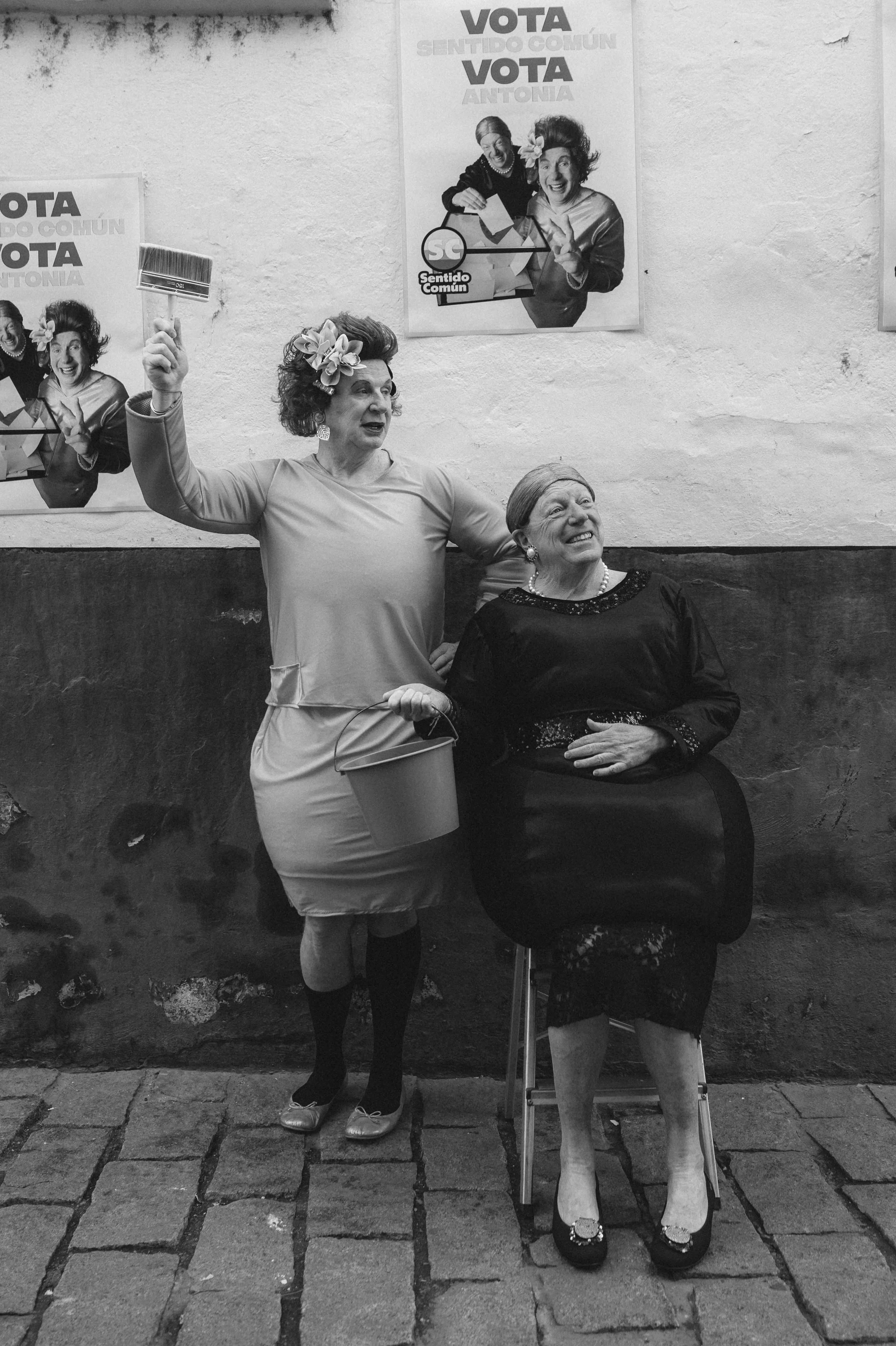 Antonia y Omaita con un cepillo en una mano y una cubeta en la otra, y otra está sentada en una silla, usando un vestido negro y tacones. Un cartel con el texto 'Vota Sentido Común' y otro con 'Vota Antonia'.