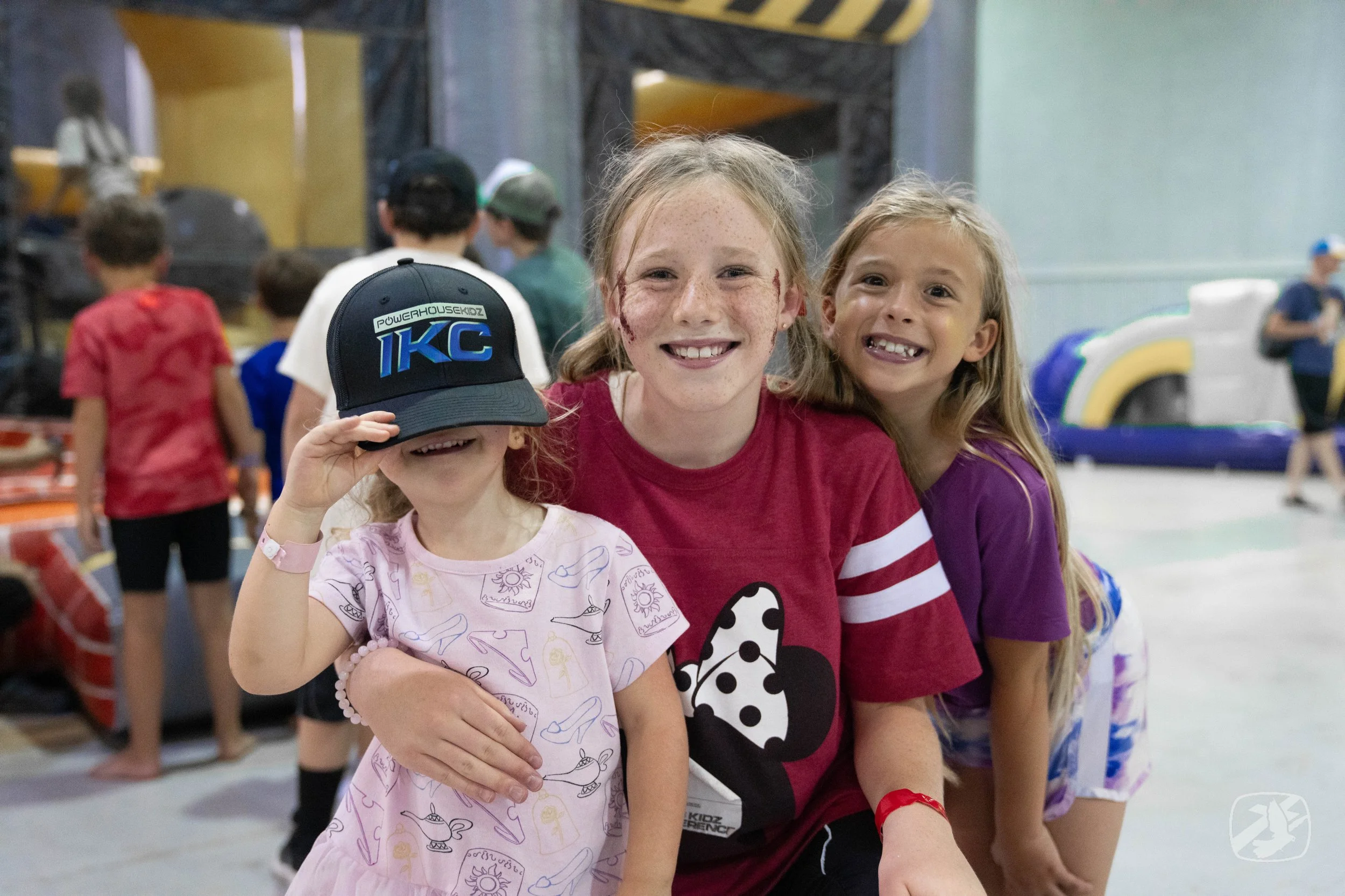 Smile of three young girls at arcade or indoor play area, with children playing in background.