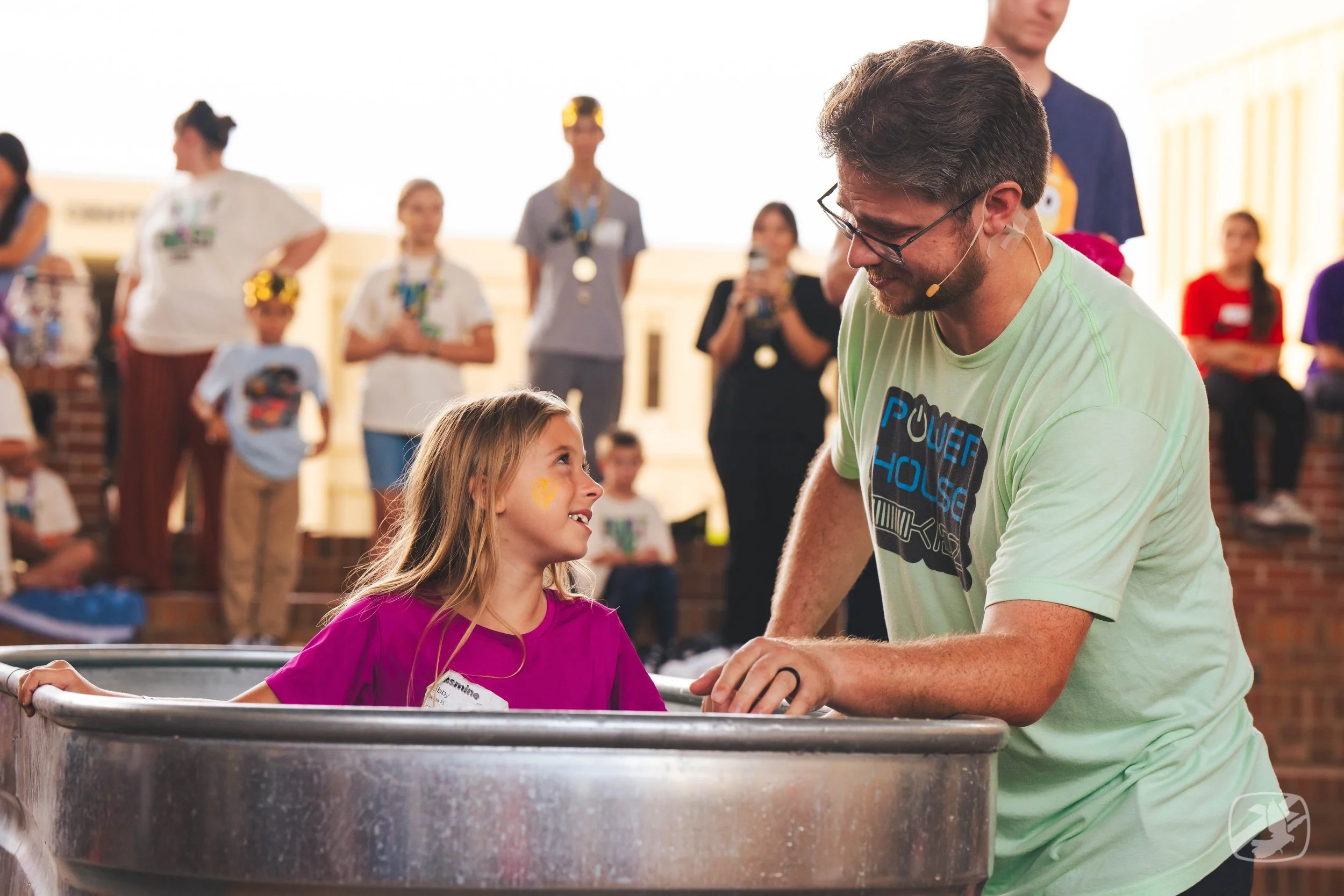 A young girl and a man are at a fair or amusement park, engaging in a dunk tank game, with people watching and waiting in the background.