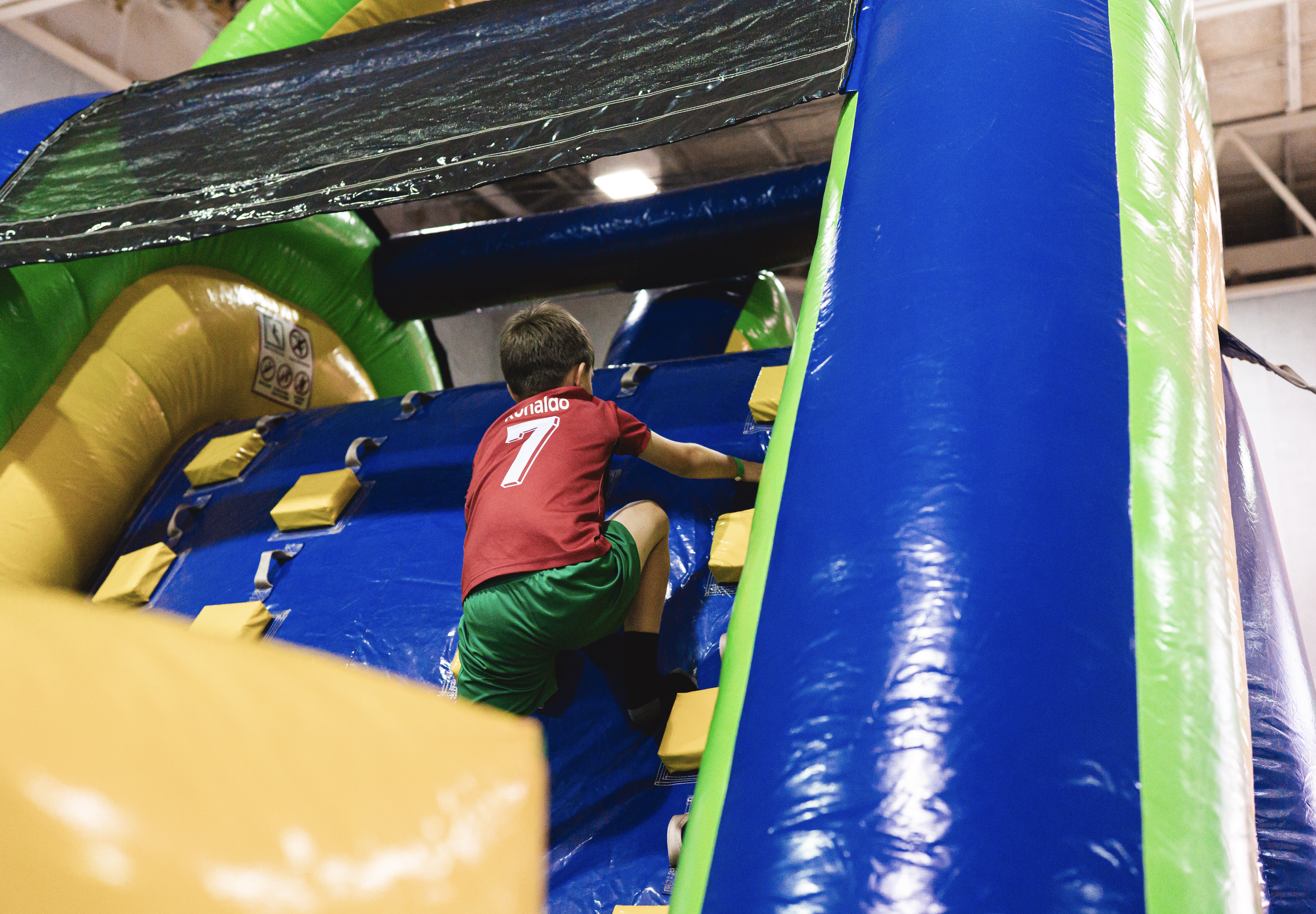 A young boy in a red shirt and green shorts climbing an indoor inflatable bounce house or obstacle course with colorful blue, green, and yellow sections.