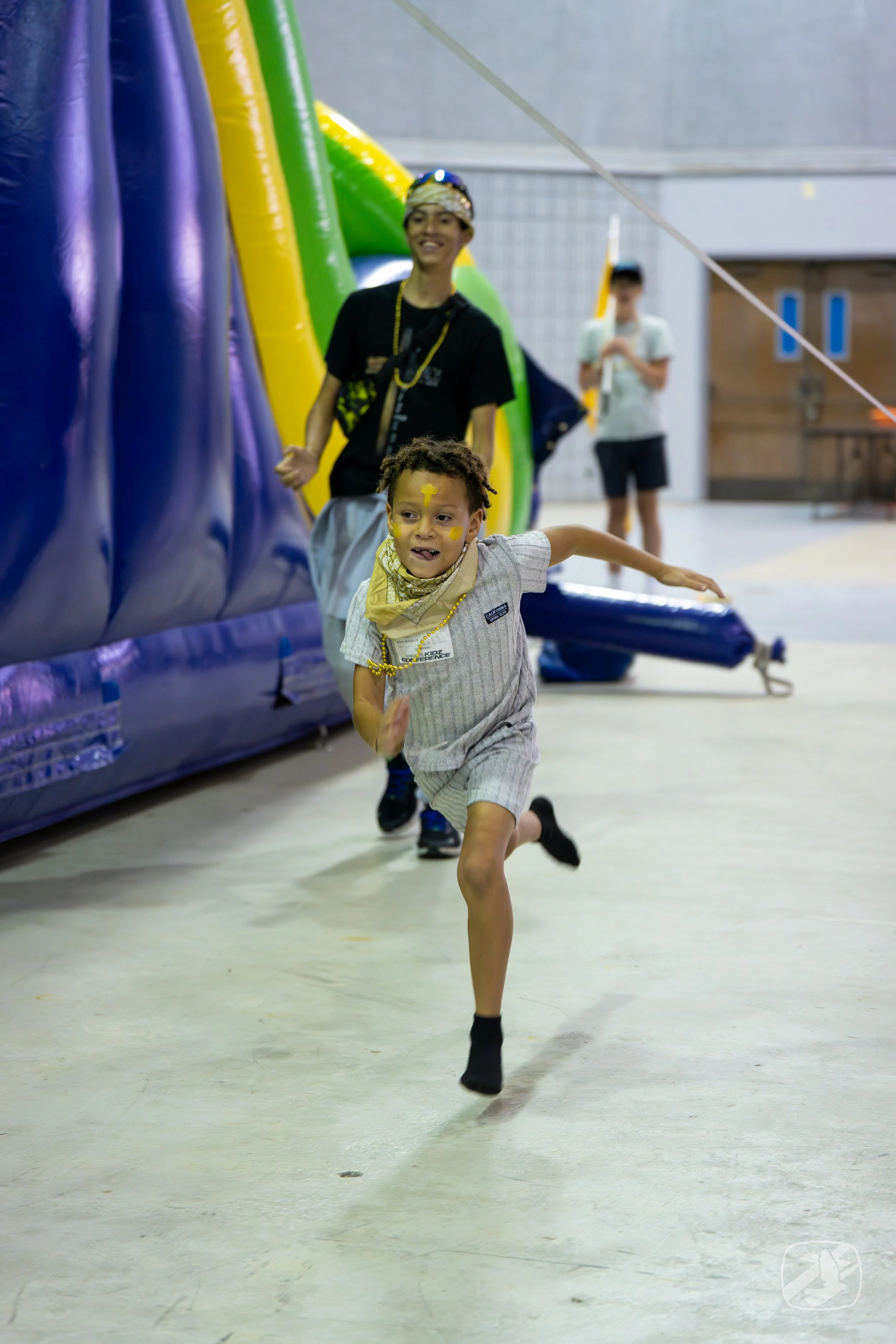 A young boy with face paint running indoors on a light-colored floor, with a smiling adult and another person in the background near an inflatable structure.