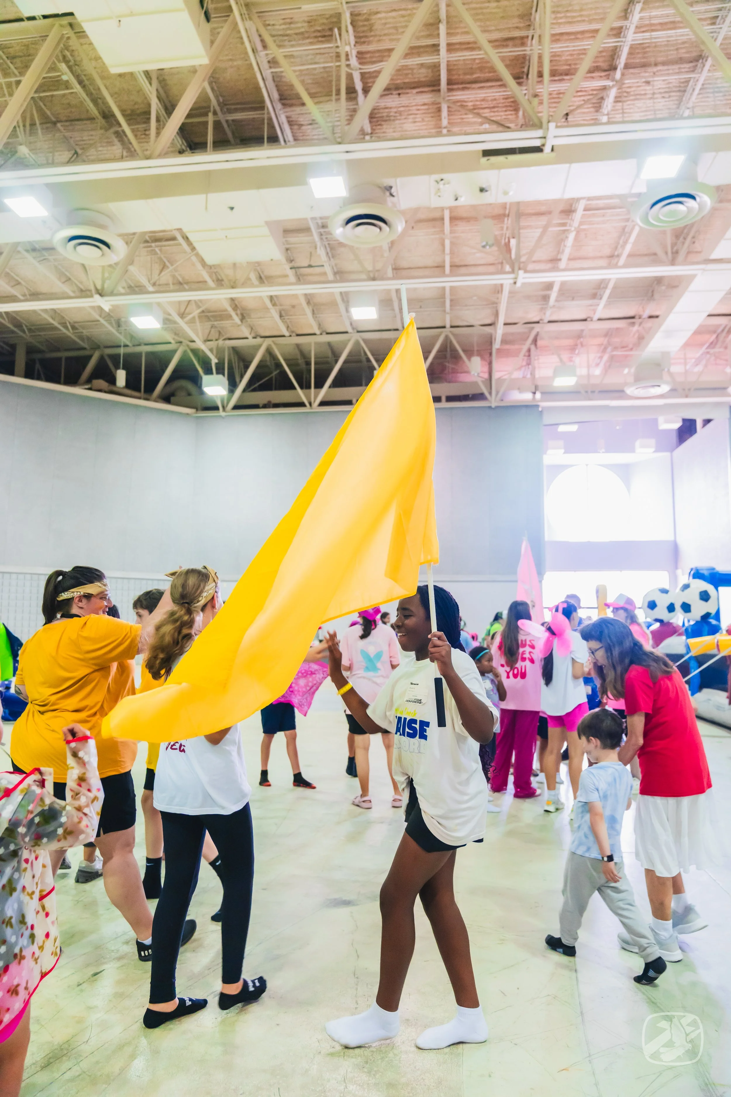 People participating in a lively indoor event, some holding yellow flags, with others wearing costumes and colorful outfits, in a gymnasium or large hall with a high ceiling.