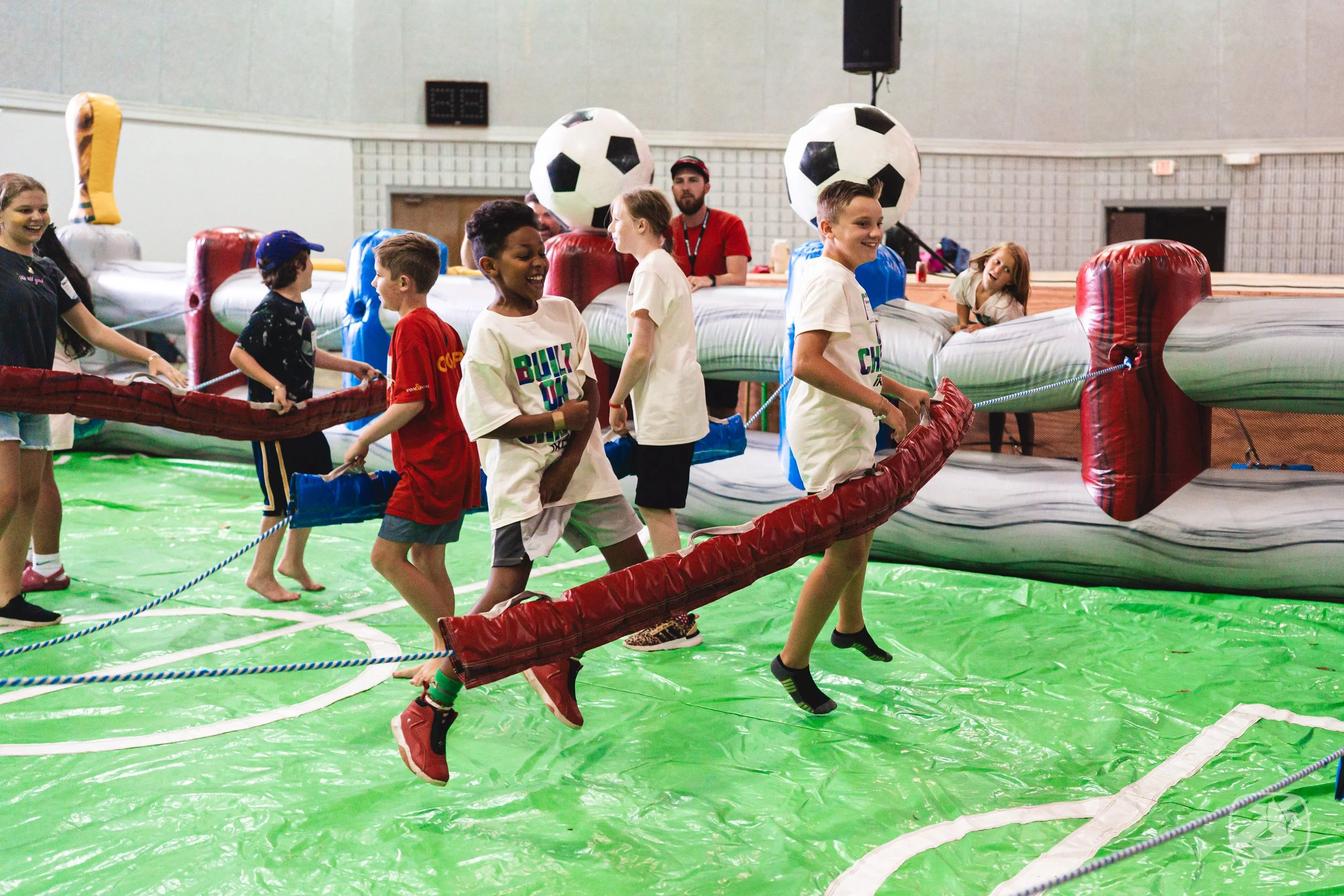 Children riding an inflatable bumper boat ride with soccer ball-shaped balloons in the background.