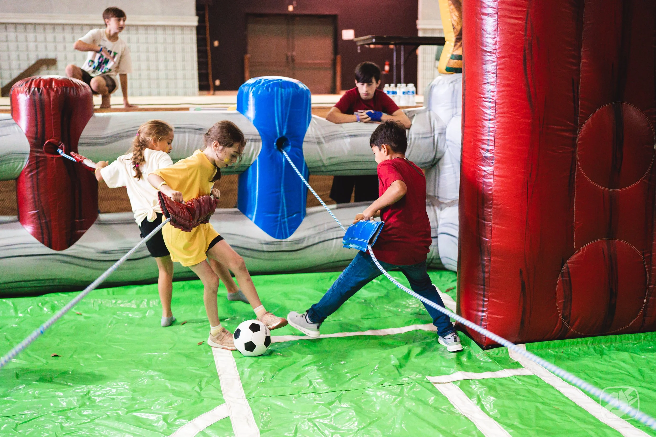 Children playing soccer inside an inflatable obstacle course with red and blue sections, on a green surface.