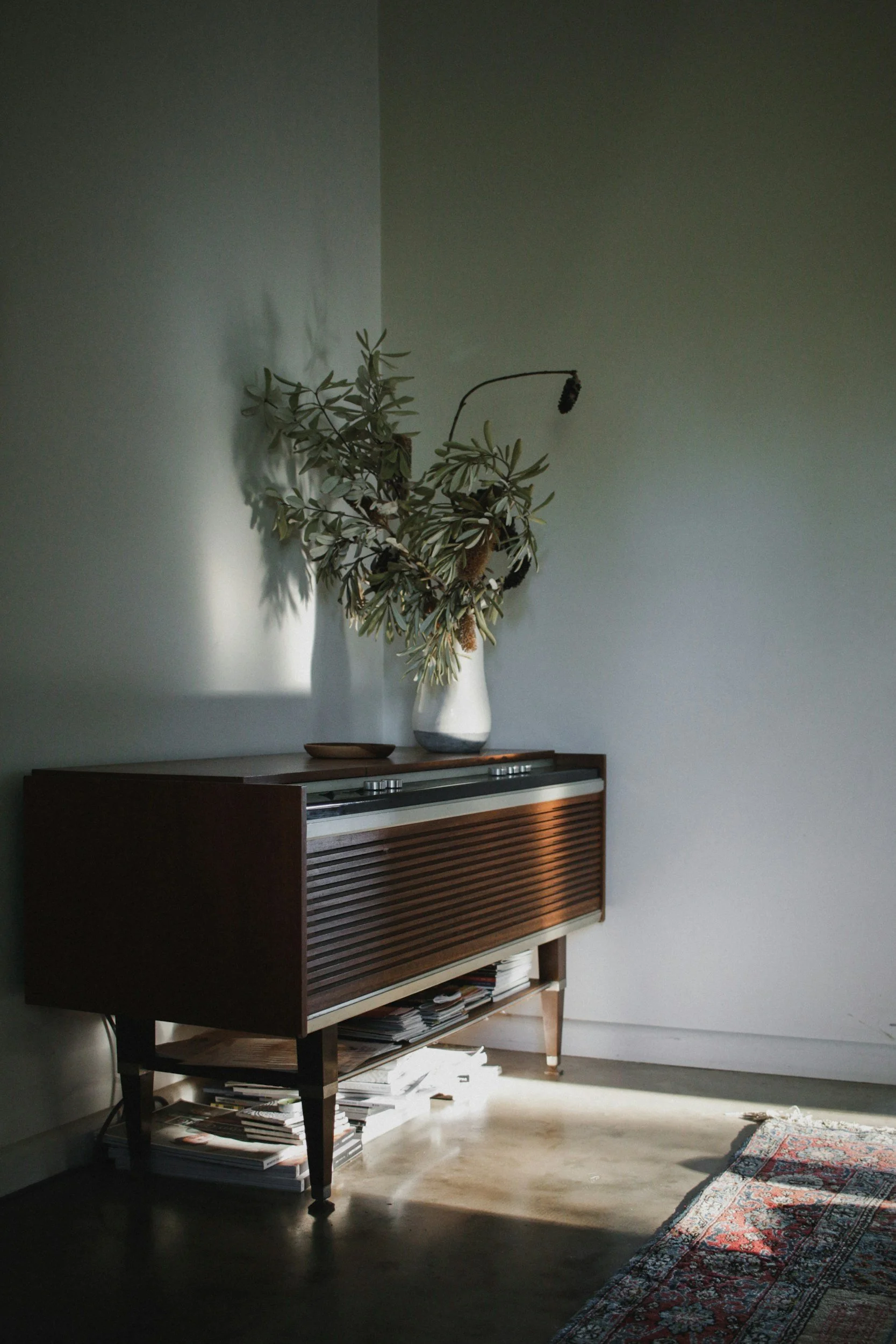 A vintage wooden sideboard with slatted doors in a room with a grey wall, decorated with a large vase of dried branches and a small decorative bowl, with sunlight casting shadows and a patterned rug on the floor.