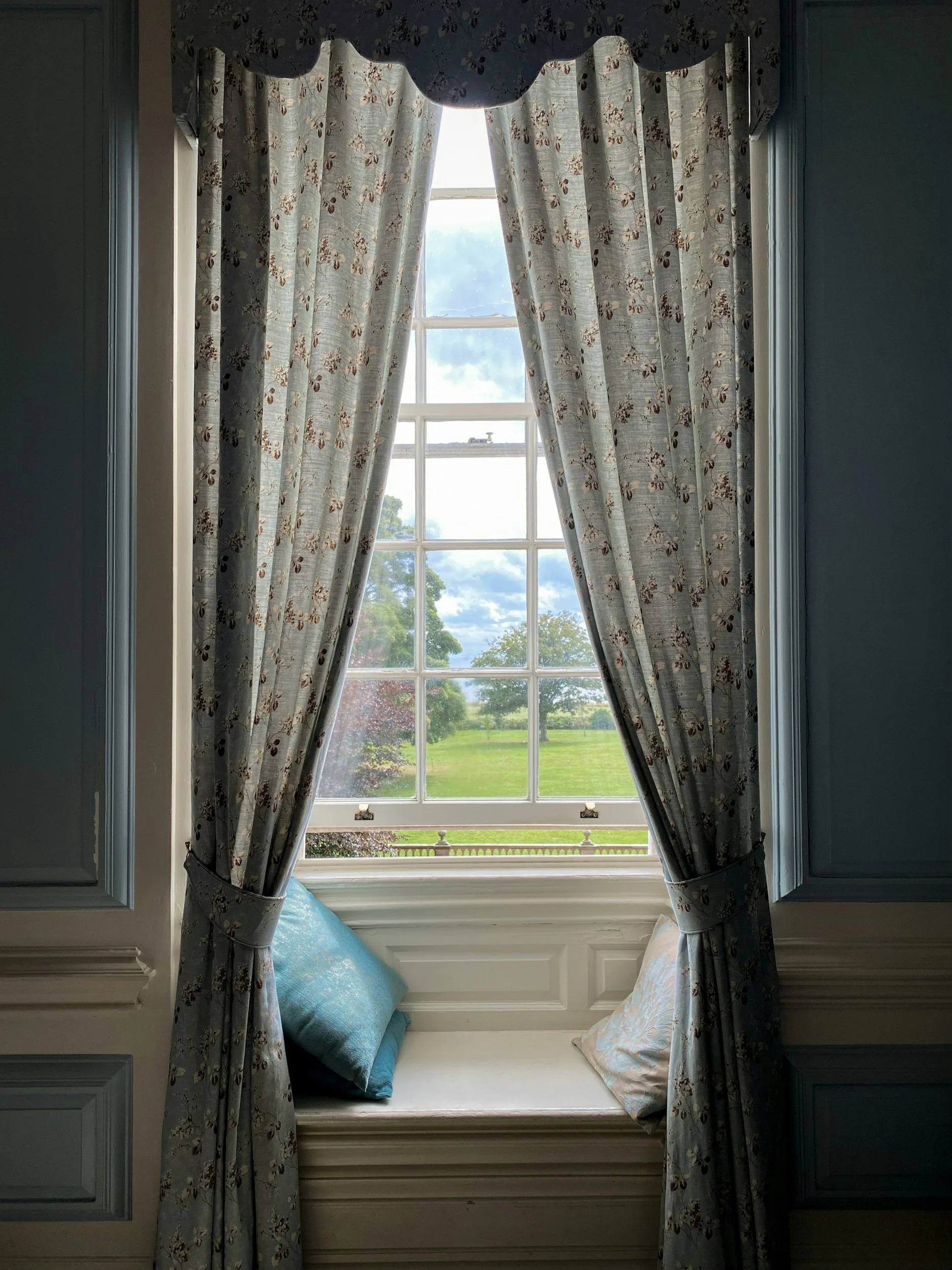 A window seat with blue pillows framed by floral curtains, showing a view of a green lawn and trees outside on a partly cloudy day.