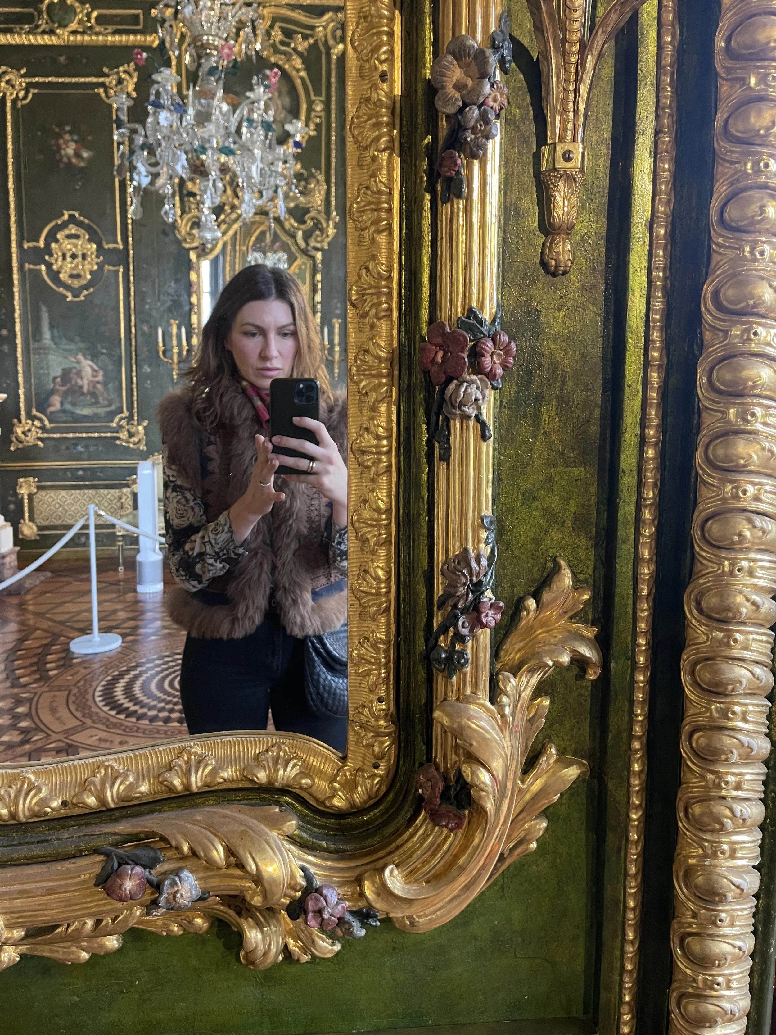 A woman taking a mirror selfie in a richly decorated room featuring gold accents, ornate carvings, floral embellishments, green walls, and a chandelier.
