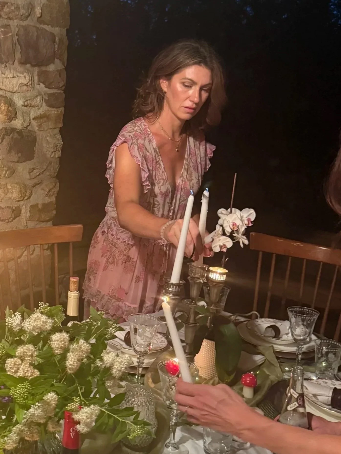 A woman in a pink floral dress stands at a decorated table, lighting candles during an evening gathering.