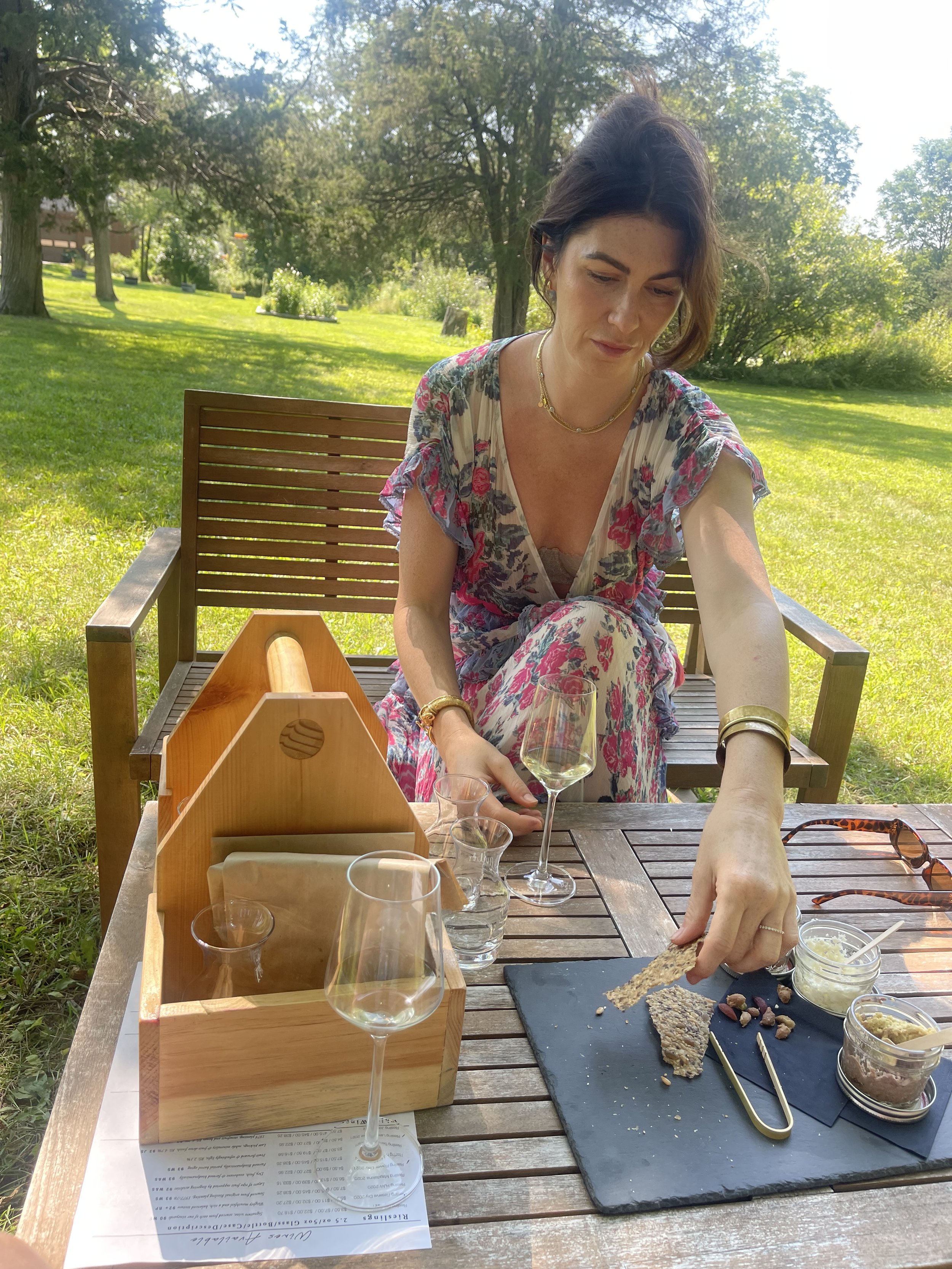 Woman in floral dress pouring cheese and snacks onto a black slate board at an outdoor table with wine glasses, small bowls, and a wooden holder on a sunny grassy area with trees in the background.
