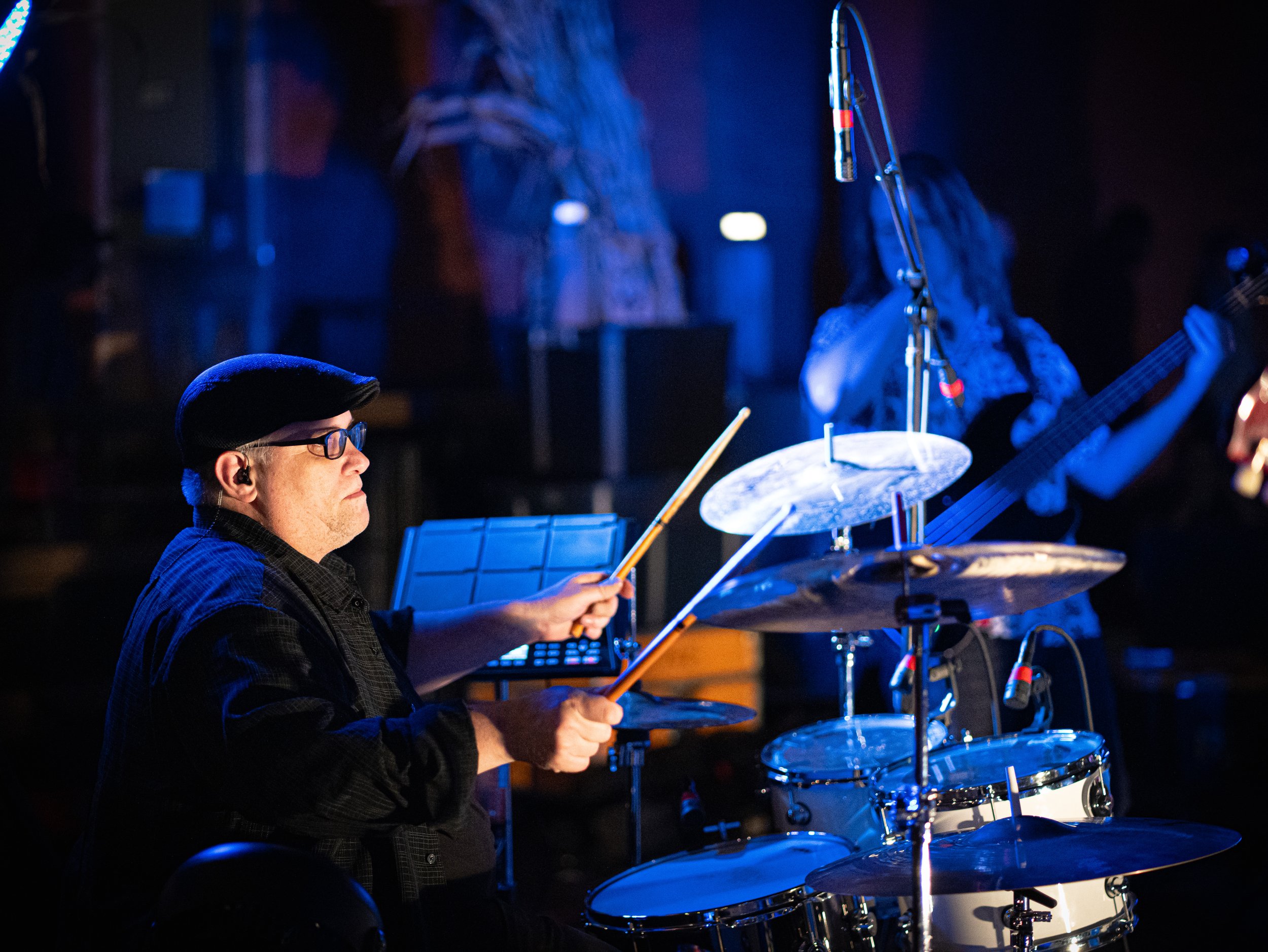 A musician playing drums on stage with a woman in the background playing guitar, both illuminated by blue stage lighting.