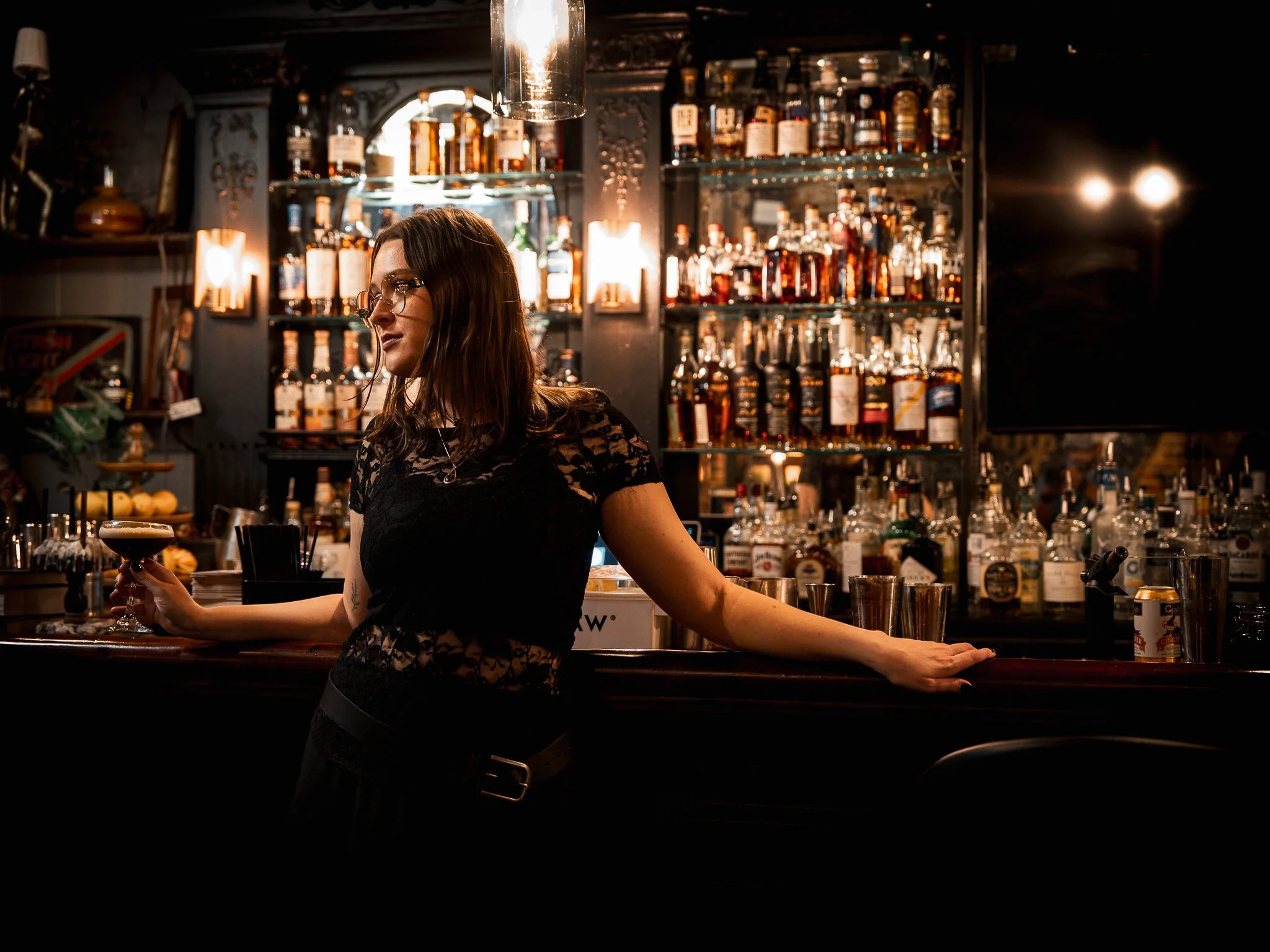 A woman bartender with glasses and long dark hair working behind a bar, holding a cocktail glass with a dark drink, in a dimly lit bar with shelves of liquor bottles in the background.