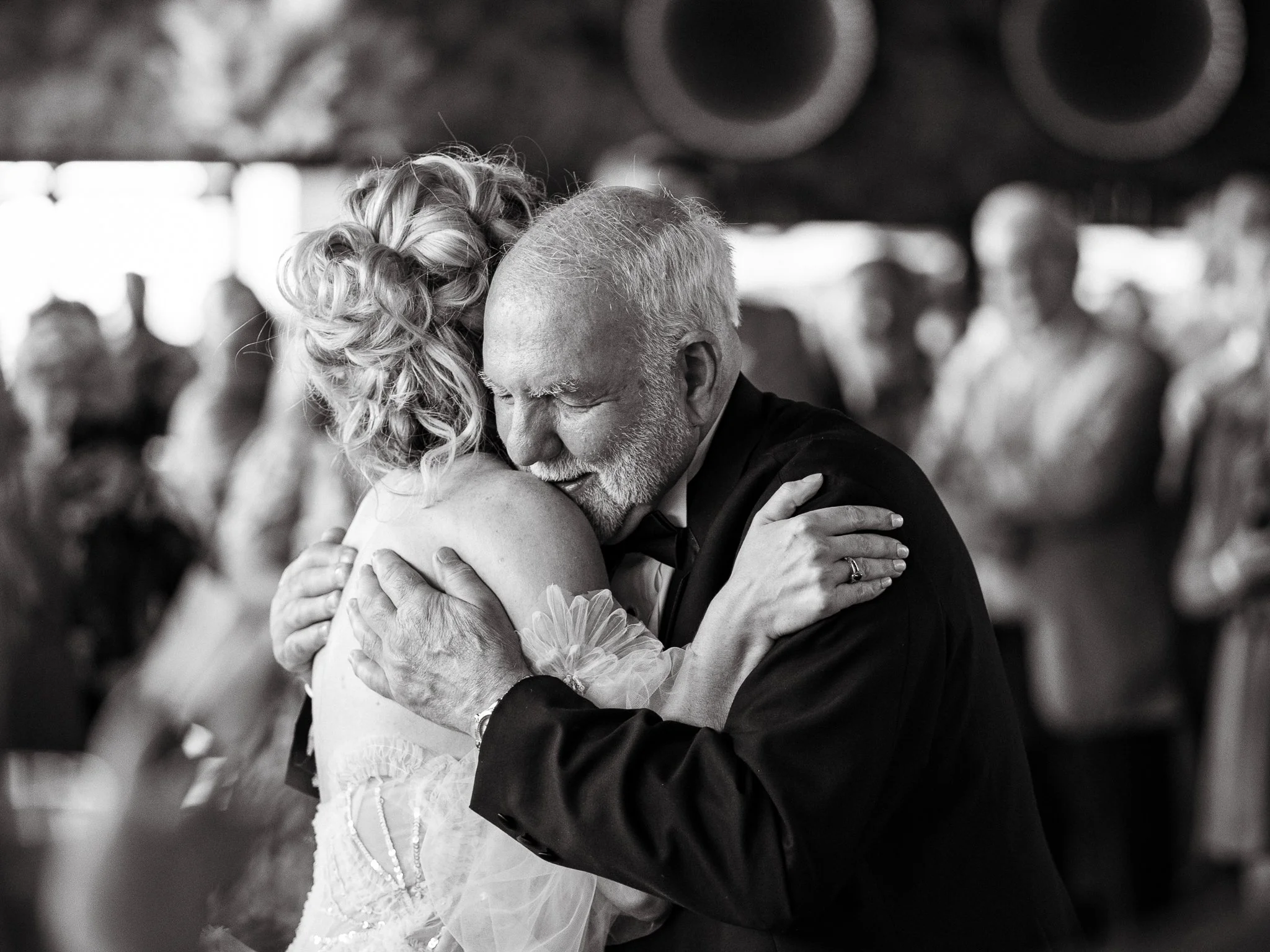 A black and white photo of a couple dancing closely at a wedding reception, with a blurred background of other guests.