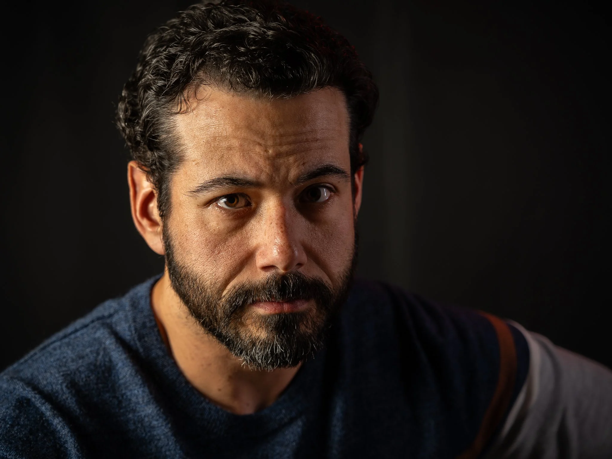 Close-up of a man's face with a beard and mustache, dark curly hair, and brown eyes, wearing a dark blue hoodie against a black background.