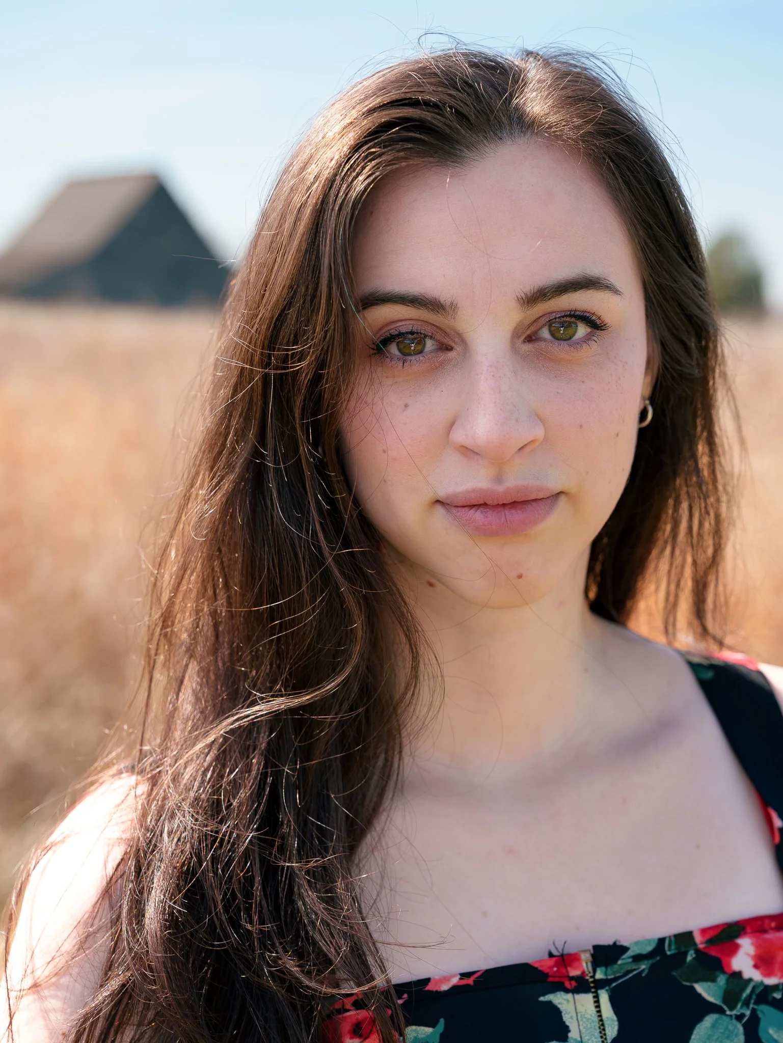 Close-up of a young woman with long brown hair, green eyes, and freckles, outdoors on a sunny day, wearing a black floral dress.