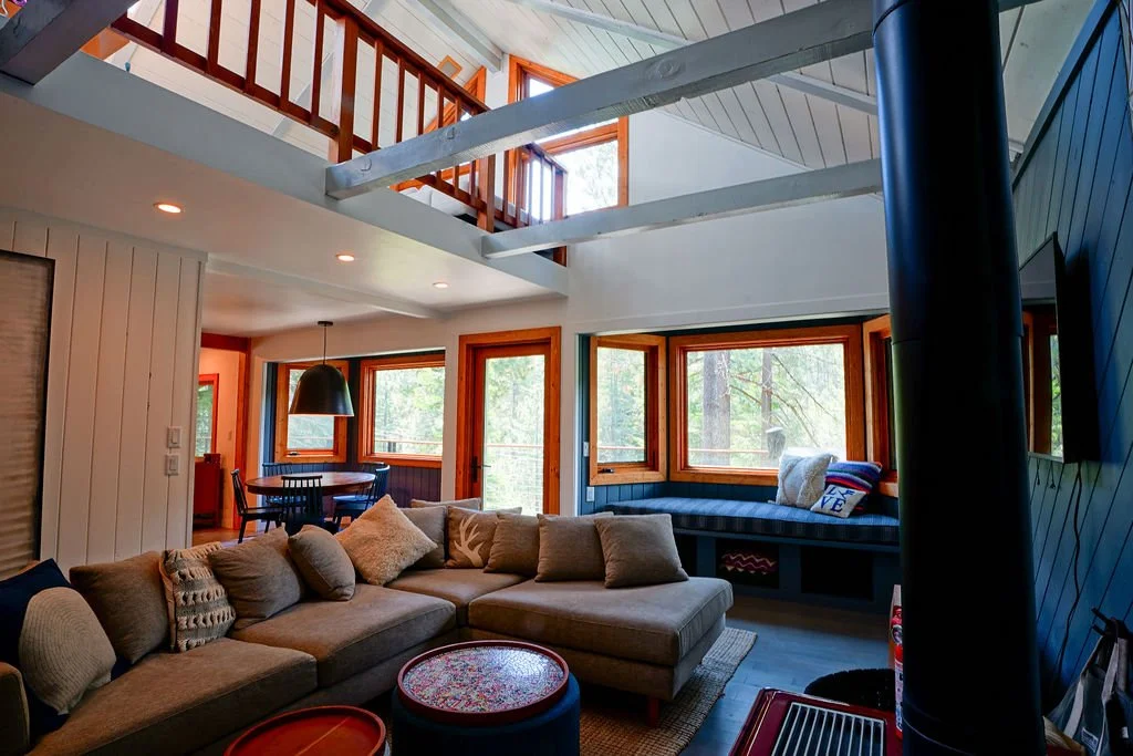 Living room with a gray L-shaped couch, freestanding wood stove, exposed beams, a loft above, and large windows filling the space with natural light.