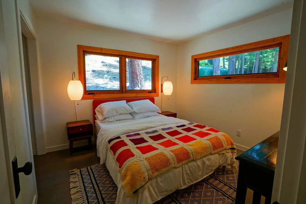 Small bedroom with white walls, wood-framed windows, dark flooring, a black and white patterned rug, and a bed with white bedding and a colorful block-pattern blanket.