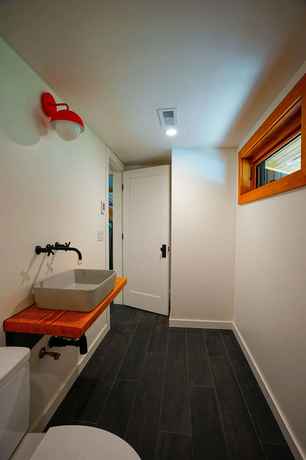 Narrow bathroom with white walls, long narrow window near ceiling, black flooring and fixtures, a floating wood vanity with gray sink basin, and a small red wall light.