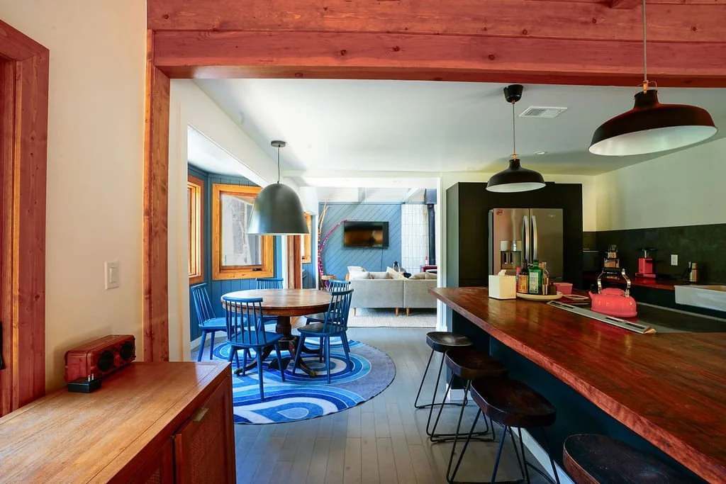 Kitchen with exposed wood beams, a large island with wood countertops and range, opening into a bright dining area with a circular table, metal pendant light, and patterned rug.