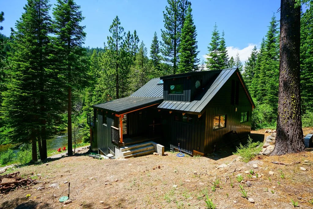 Black-sided cabin-style home with a black metal roof, surrounded by trees beside a river with two red Adirondack chairs down by the water.