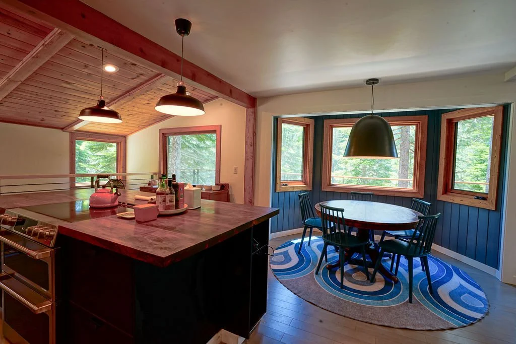 Dining area with a circular table, blue wooden chairs, a large green pendant light, and windows beside an open kitchen with a large island.