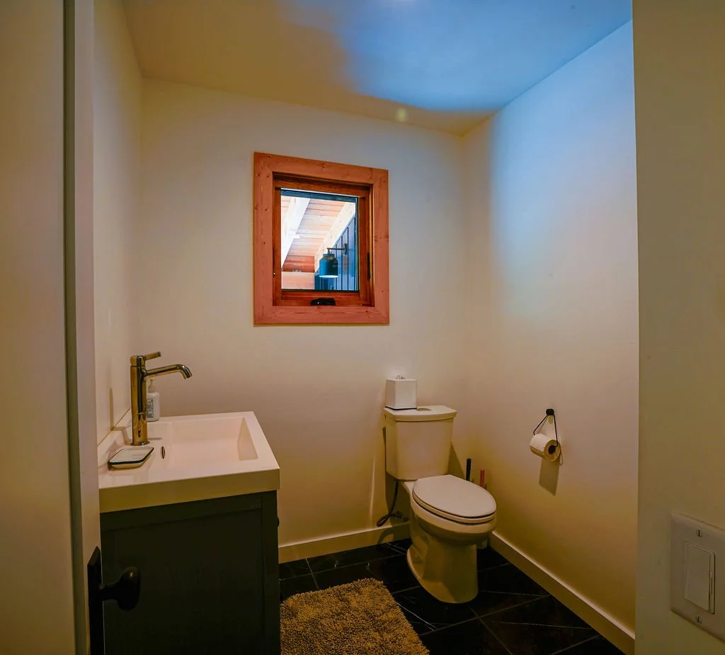 Small bathroom with white walls, dark flooring, a dark vanity with a white countertop, a toilet, and a small square window.
