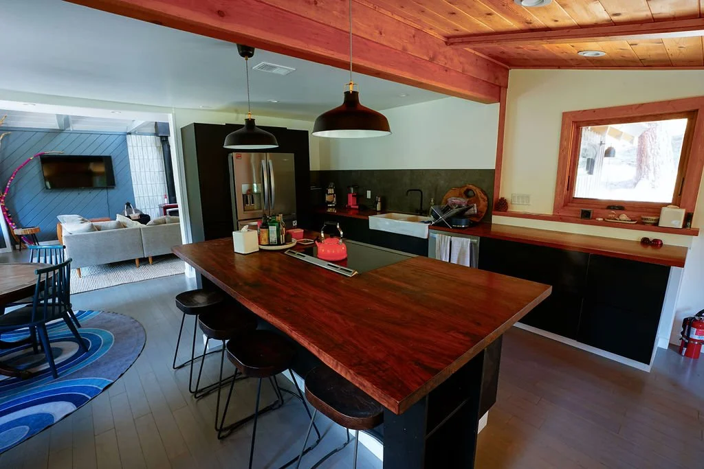 Kitchen with exposed wood beams, a large island with wood countertops and range, black pendant lights, and an open view into the bright dining area and living room.