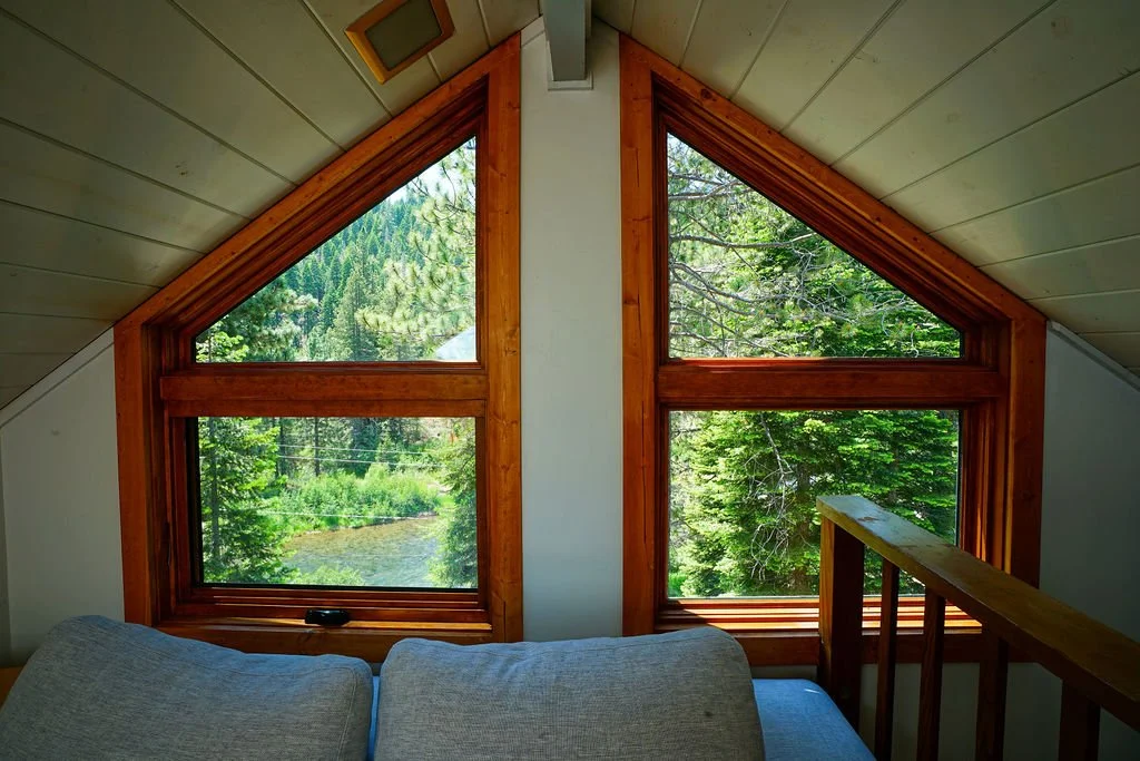 Triangular windows beneath a peaked white ceiling overlooking a wooded setting and river.
