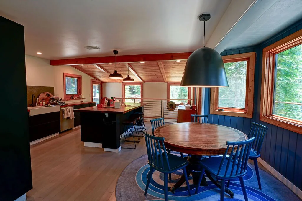 Bright dining area with circular table and blue wooden chairs and large green pendant light with large windows set into a popped out blue accent wall and an open view into the kitchen.