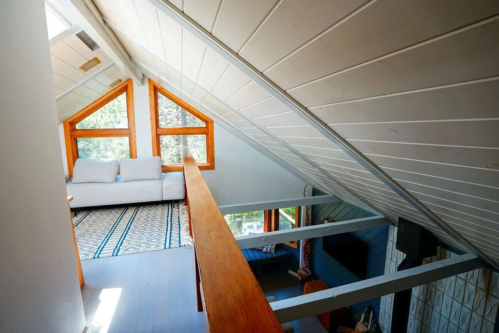 Bright loft with a blue patterned rug, wooden railing, exposed beams overlooking the living room, white ceiling, and triangular windows.
