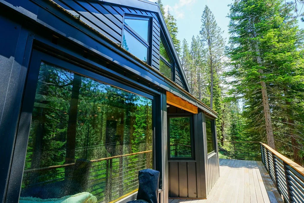 Black-sided cabin-style home with a peaked roof, large windows looking over large wood deck, modern metal railing, and a wooded natural setting.