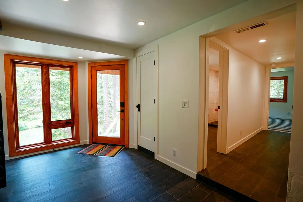 Bright entryway with a red front door, tall windows, dark wood flooring, white walls, a colorful rainbow rug, and a hallway leading to additional rooms.
