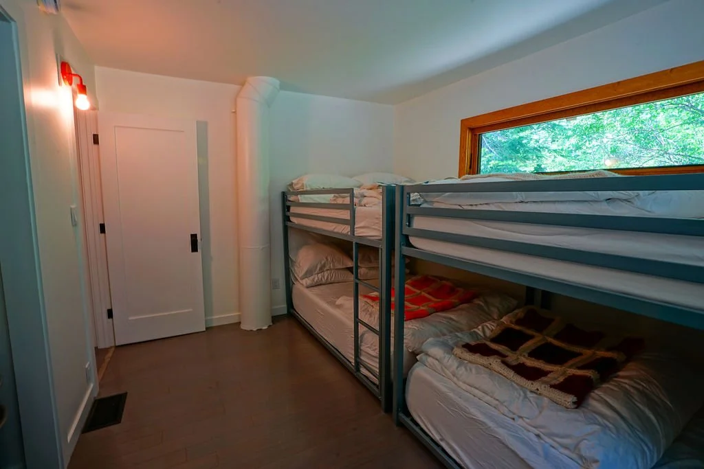 Bunk room with two side-by-side bunk beds, white walls, dark wood flooring, a narrow window above the bunks, and a doorway leading from the room.