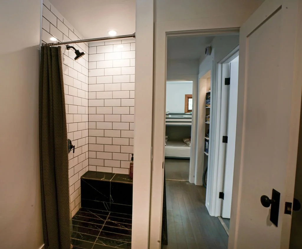 Small bathroom with white subway tile walls, a shower stall with a black curtain, black subway tile flooring and bench, and a view toward the hallway leading to the bedroom.