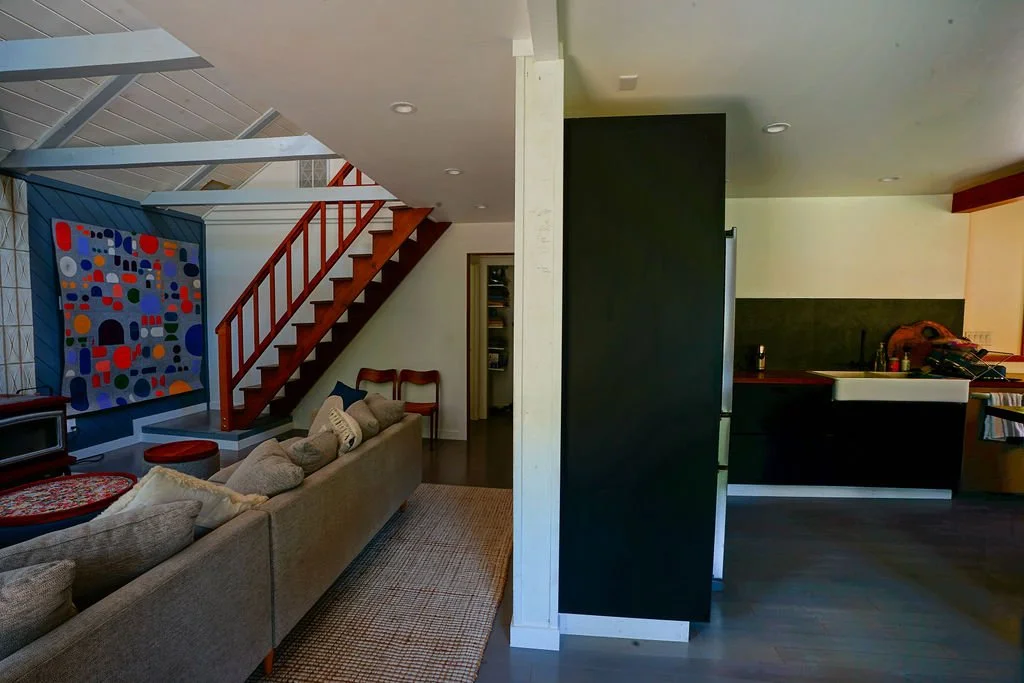 Living room with a large tan couch, stairs leading to the upper level beside a dividing wall, and an open view into the kitchen on the right.