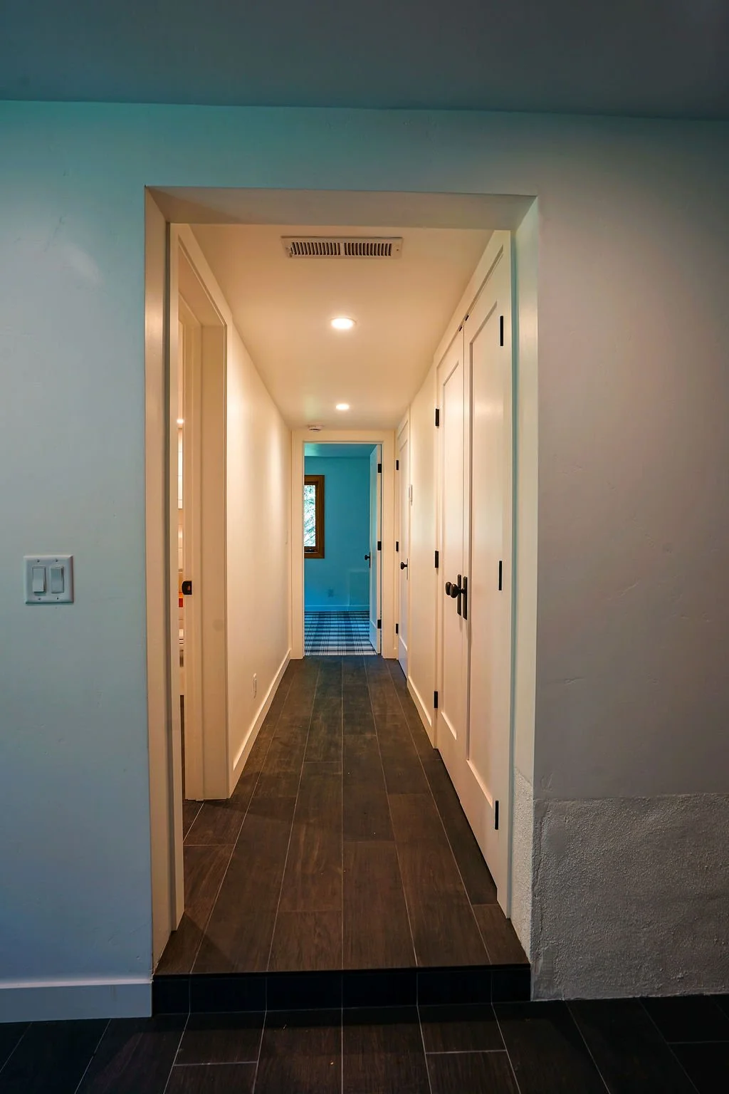 Long hallway with white walls, dark wood flooring, and lighting leading to an adjoining room.