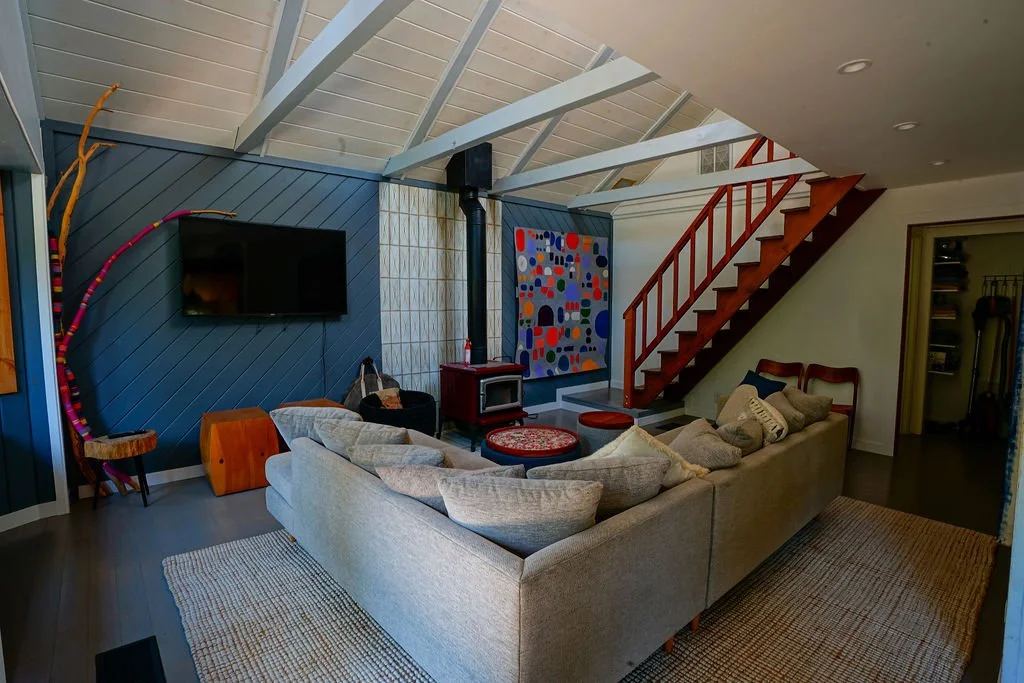 Living room with gray flooring, a large tan rug and couch, blue accent wall, white tiled wall behind a freestanding wood stove, and wooden stairs leading to the upper level.