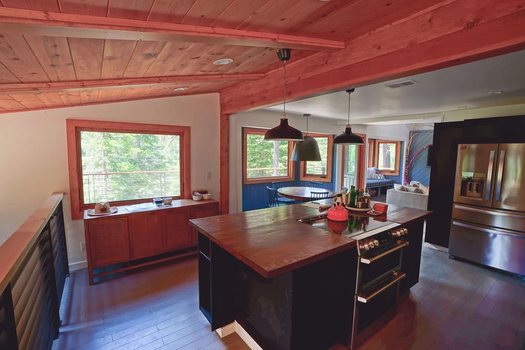 Kitchen with exposed wood beams, wood countertops, dark cabinetry, a stainless steel refrigerator, a large island with range, and a large window.